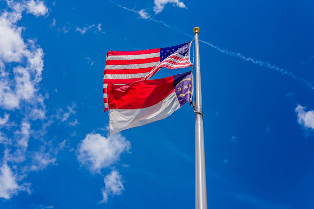 American and North Carolina state flags waving against a clear blue sky with scattered clouds.